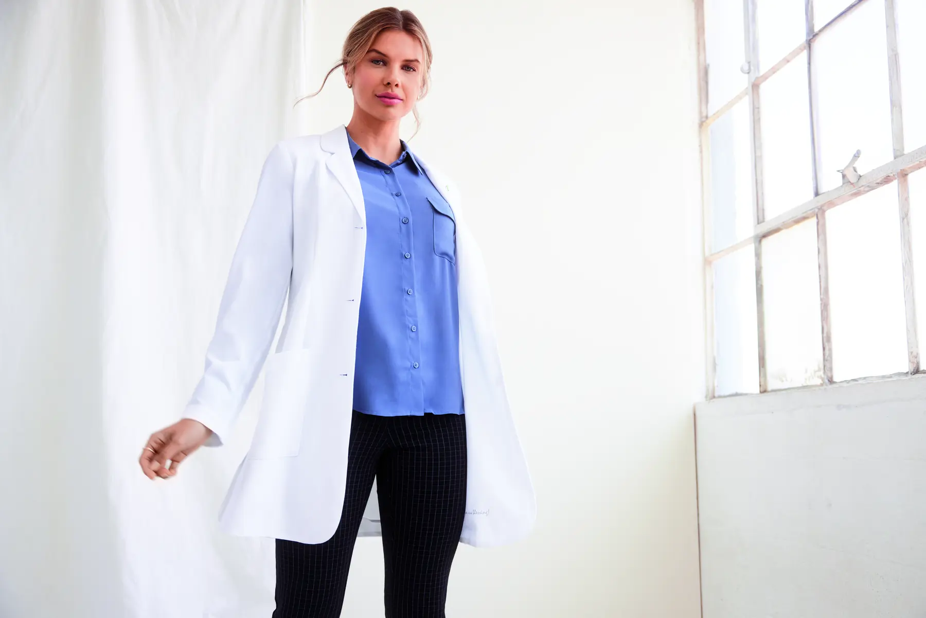 Medical professional wearing a white lab coat over business attire, standing confidently in a bright room.